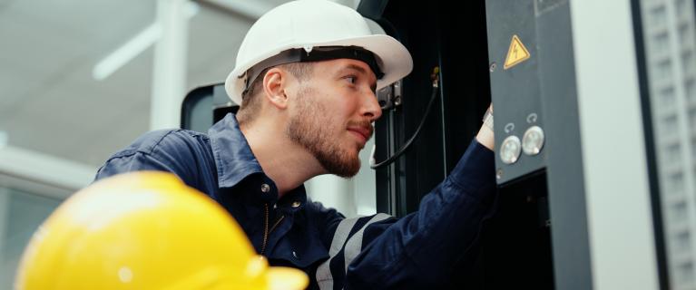 Installateure Zwei Techniker mit Schutzhelmen arbeiten an einem geöffneten Schaltschrank; einer prüft innen die Komponenten, der andere bedient mit Werkzeug den unteren Bereich.