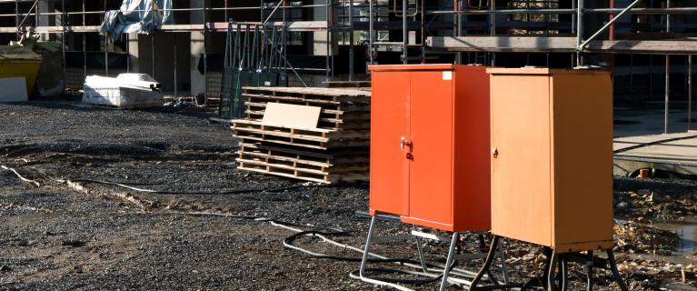 Zwei Baustromverteiler mit Kabeln auf Schotter vor einem eingerüsteten Gebäude.