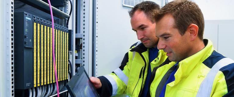 Zwei Techniker in Warnjacken arbeiten mit Laptop an einem Schaltschrank mit Modulen.