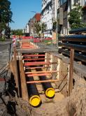 Baustelle in einer Stadtstraße: ein offener Rohrgraben mit verlegten Leitungen und daneben gestapelte, schwarz isolierte Rohre mit gelben Endkappen.