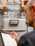 Mann liest einen Gaszähler an der Wand ab und notiert Werte auf einem Tablet; darüber verlaufen Kupferrohre mit zwei gelben Absperrhähnen.
