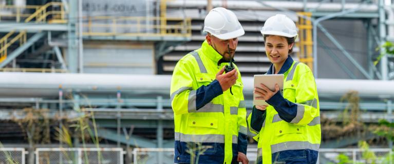 Zwei Personen in Warnjacken und Helmen stehen vor einer Industrieanlage; eine nutzt Funkgerät, die andere hält ein Tablet.