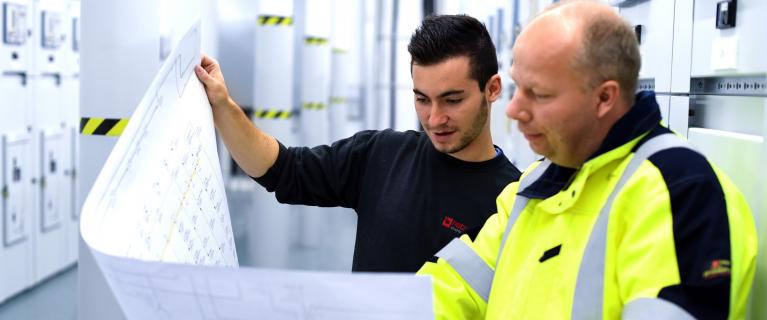 Zwei Techniker in einem Schaltraum prüfen gemeinsam eine große technische Planzeichnung.