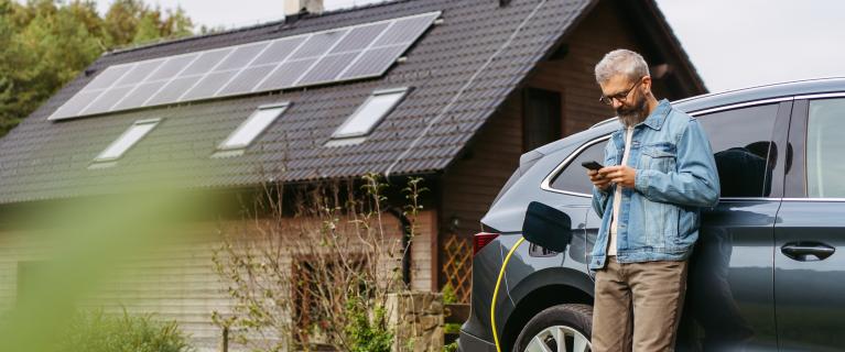 Mann steht neben einem Elektroauto beim Laden und schaut auf sein Smartphone; im Hintergrund ein Haus mit Solarpanels auf dem Dach.