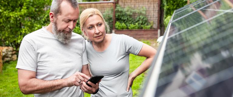 Mann und Frau stehen neben Solarmodule im Freien. Mann schaut auf ein Smartphone.