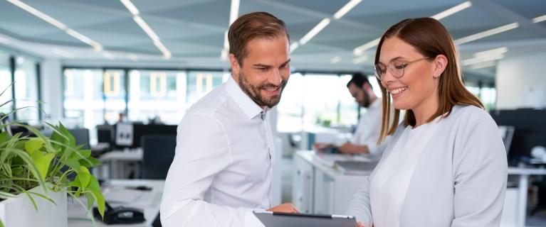 Mann und Frau stehen an einem Tresen im Büro und schauen lächelnd auf ein Tablet.
