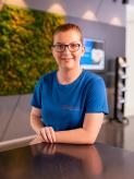 Frau mit Brille in blauem T-Shirt steht an einer Theke in einem modernen Empfangsbereich; im Hintergrund eine grüne Pflanzenwand.