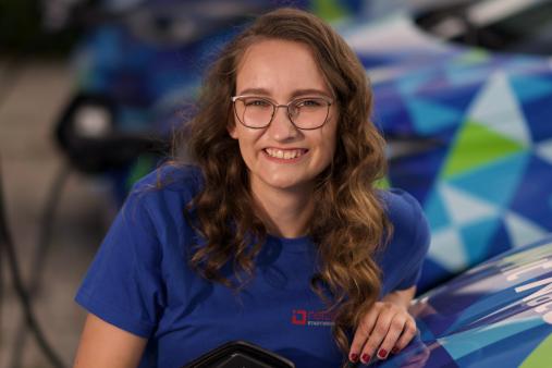 Lächelnde Frau mit Brille in blauem Shirt hält ein Ladekabel an einem E-Auto; im Hintergrund weitere Fahrzeuge.