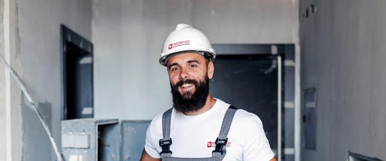 Techniker mit weißem Schutzhelm und Arbeitslatzhose steht in einem Rohbauflur, lächelt in die Kamera und hält ein Tablet in der Hand.