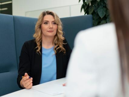 Eine Frau sitzt in einer gepolsterten Sitznische an einem Tisch und spricht mit einer unscharf im Vordergrund sichtbaren Person.