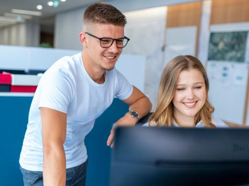 Mann stehend und Frau am Arbeitsplatz sitzend schauen gemeinsam auf einen Monitor.