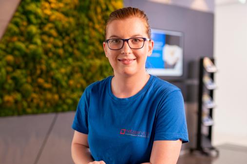 Frau mit Brille in blauem T-Shirt steht an einer Theke in einem modernen Empfangsbereich; im Hintergrund eine grüne Pflanzenwand.