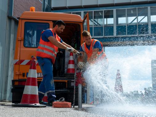 Netzservice Unternehmen Zwei Arbeiter in Warnwesten öffnen einen Hydranten auf der Straße; Wasser spritzt heraus, daneben steht ein orangefarbener Servicewagen und Verkehrskegel sichern die Stelle.