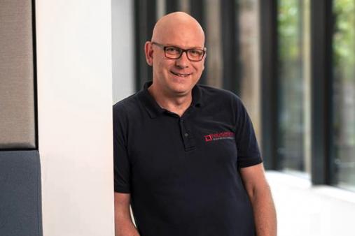 Mann mit Brille lehnt lächelnd an einer Wand in einem modernen Büroflur mit großen Fenstern.