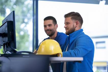 Zwei Männer stehen am Arbeitsplatz vor einem Monitor; ein gelber Schutzhelm liegt im Vordergrund auf dem Tisch.