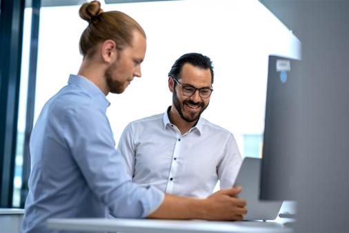 Zwei Männer stehen im Büro an einem Schreibtisch und schauen gemeinsam auf einen Monitor; einer lächelt.
