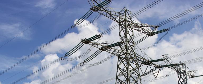 Hochspannungsmasten und Stromleitungen vor einem Kraftwerksgelände mit Schornsteinen.