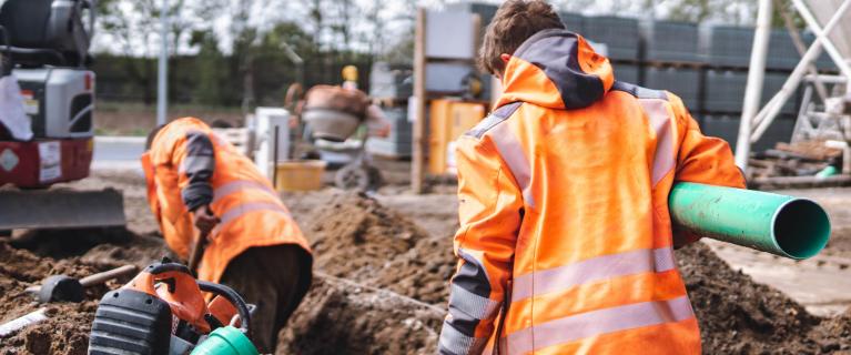Zwei Bauarbeiter in Warnkleidung verlegen ein Rohr in einer Baugrube.