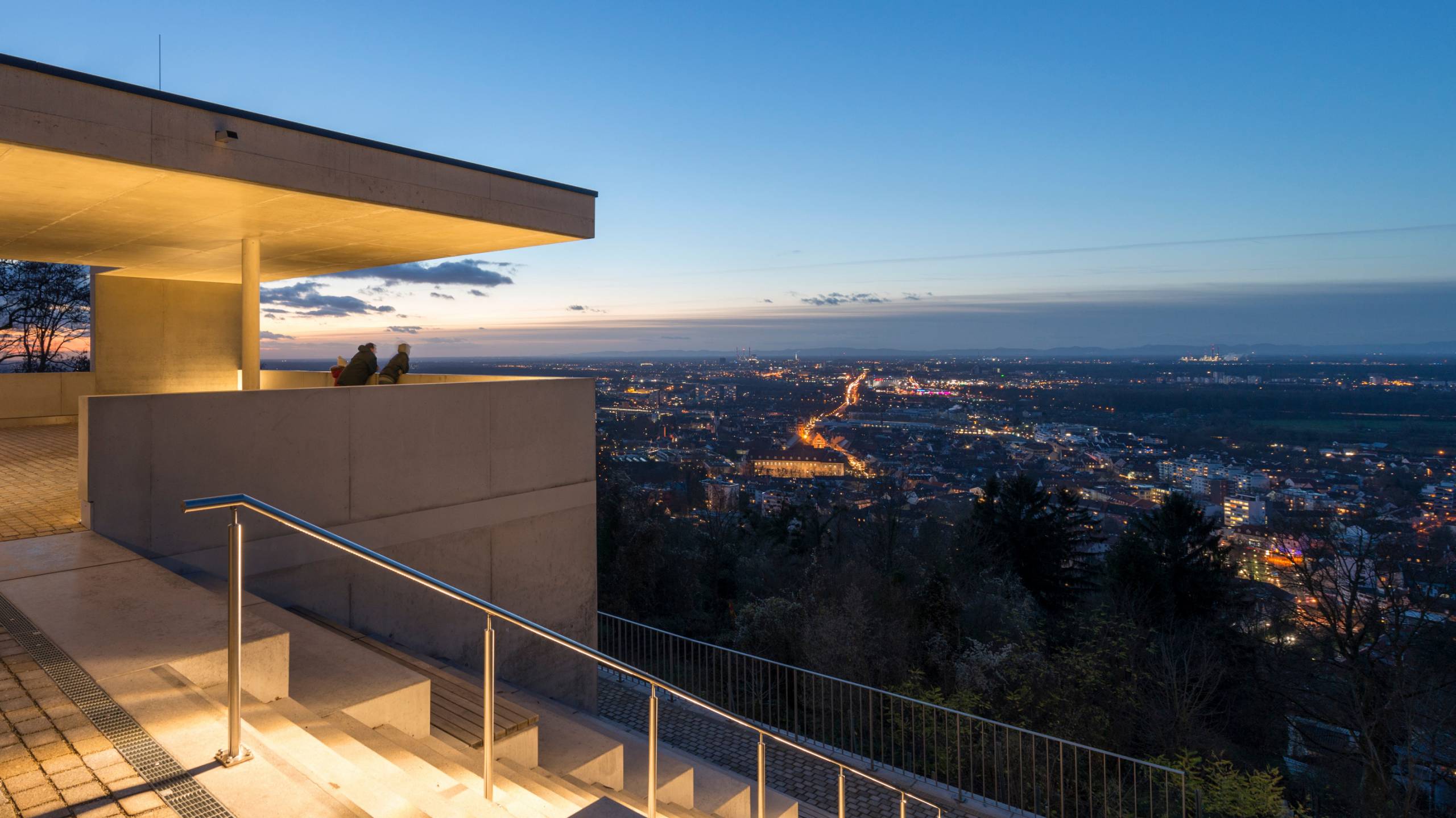 Zwei Personen stehen auf einer überdachten Aussichtsplattform und blicken bei Dämmerung über die beleuchtete Stadt Karlsruhe.