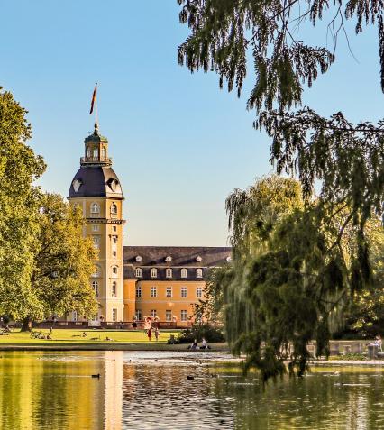 Karlsruhe Nachhaltikeit Parklandschaft mit See und vielen Bäumen. Im Hintergrund steht das Karlsruher Schloss, am Ufer sitzen und spazieren Menschen.