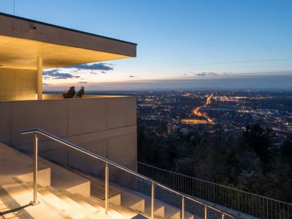 Stadtwerke Karlsruhe Netzservice GmbH Zwei Personen stehen auf einer überdachten Aussichtsplattform und blicken bei Dämmerung über die beleuchtete Stadt Karlsruhe.