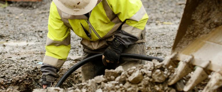 Bauarbeiter in Warnjacke und Helm arbeitet in einem Graben und verlegt ein schwarzes Kabel. Rechts ist die Schaufel eines Baggers zu sehen.
