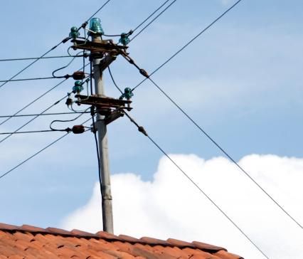 Stromleitungen an einem Mast verlaufen über ein Ziegeldach mit gemauertem Schornstein vor blauem Himmel.
