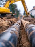 Baumaßnahmen Zwei große Rohre liegen in einem frisch ausgehobenen Graben; im Hintergrund arbeitet ein gelber Bagger auf einer Baustelle an einer Wohnstraße.