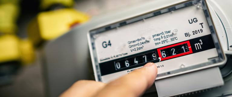 Nahaufnahme eines Gaszählers (G4) mit mechanischem Zählwerk; ein Finger zeigt auf den abgelesenen Verbrauch in Kubikmetern, im Hintergrund unscharfe gelbe Gasleitungen.